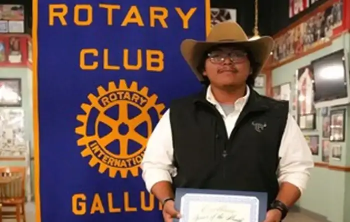 Joshua Dagen wearing a cowboy hat, glasses, a white shirt and a black vest stands indoors holding a framed certificate. Behind him is a blue banner reading "Rotary Club Gallup" with the Rotary International gear logo.