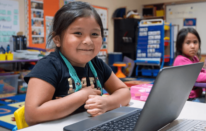 A elementary school student smiles while sitting at her computer at her desk.