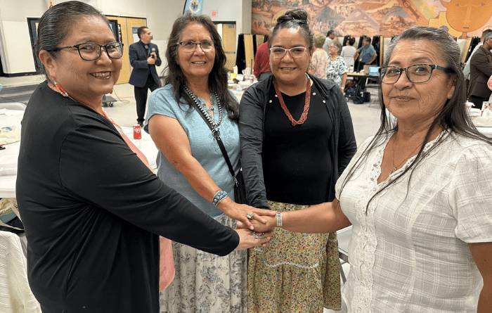 Four women stand together at a school board work session, smiling and placing their hands together in a gesture of unity.
