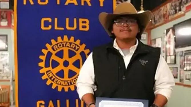 Joshua Dagen wearing a cowboy hat, glasses, a white shirt and a black vest stands indoors holding a framed certificate. Behind him is a blue banner reading "Rotary Club Gallup" with the Rotary International gear logo.