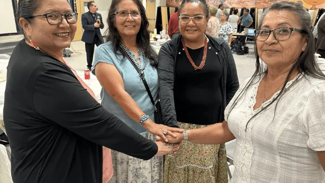 Four women stand together at a school board work session, smiling and placing their hands together in a gesture of unity.